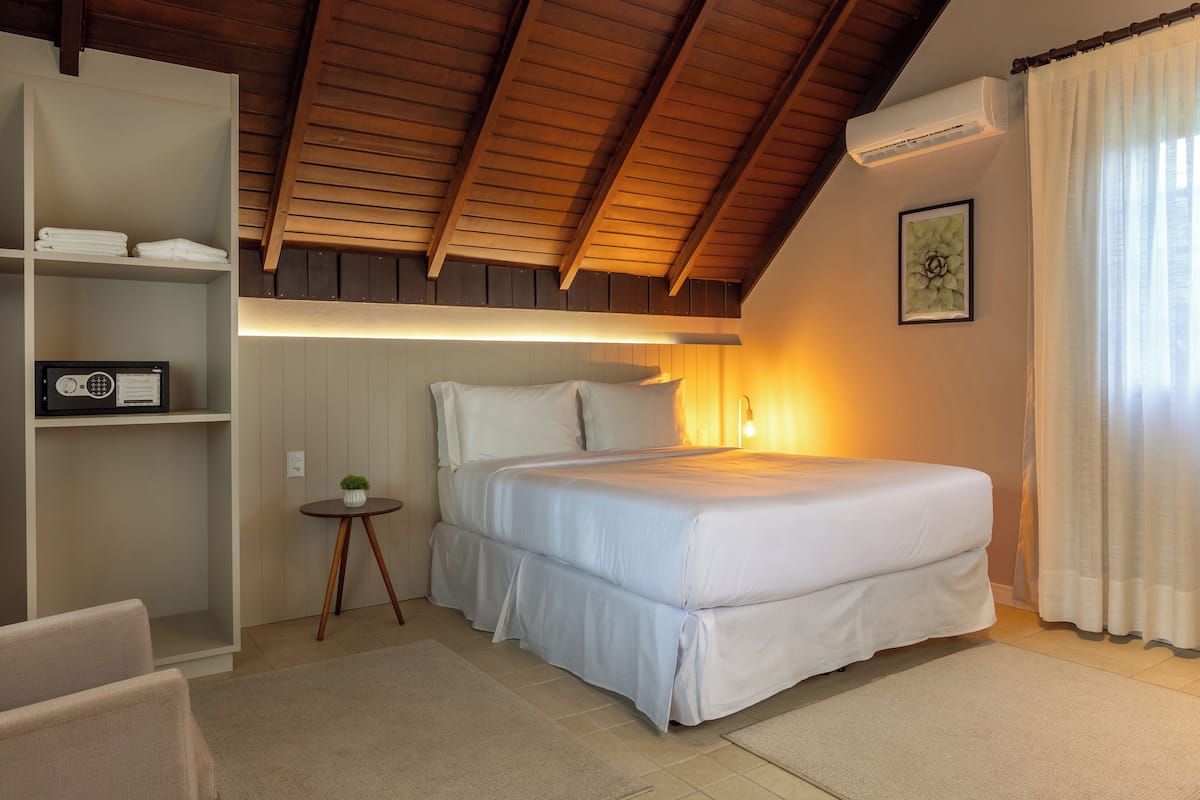 Um quarto com uma cama, uma estante com toalhas, uma mesinha com uma planta e um quadro na parede.