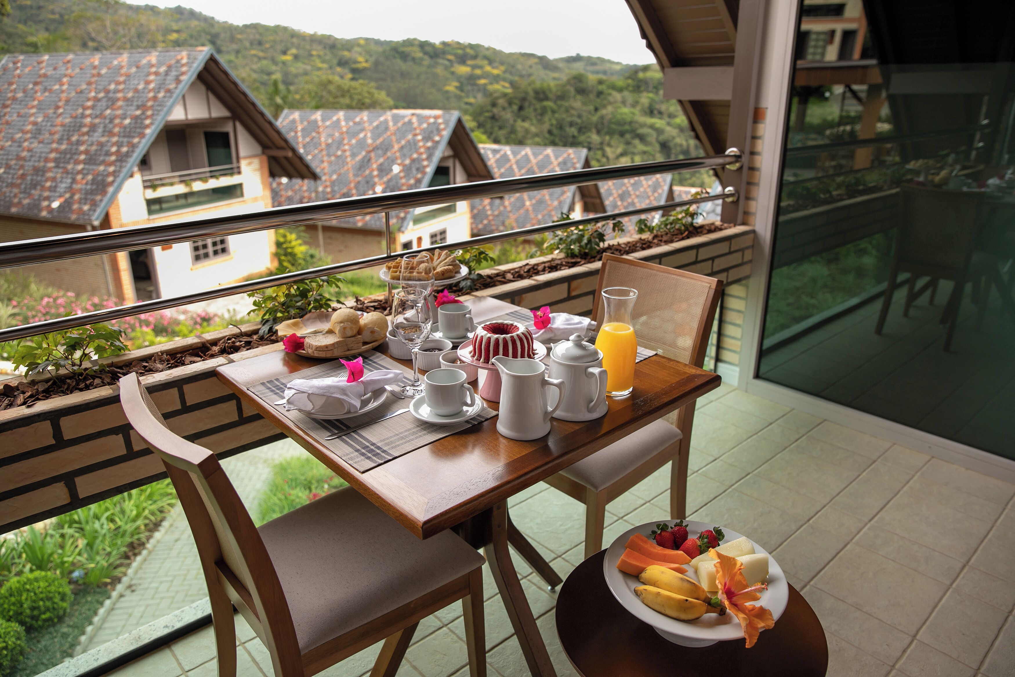 Un balcón con una mesa de desayuno, sillas y vistas a casas y vegetación.
