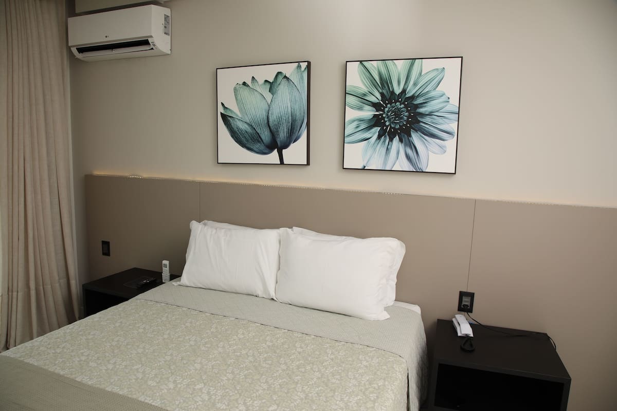 Quarto de hotel com cama, duas mesas de cabeceira, ar condicionado e dois quadros florais na parede.