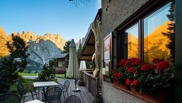 An outdoor seating area with tables and chairs, a wooden deck, and a mountain backdrop. - Rifugio La Montanara (Molveno)