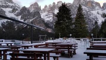 Una terraza con mesas y bancos de picnic de madera, con vistas a un paisaje montañoso con picos nevados y árboles.