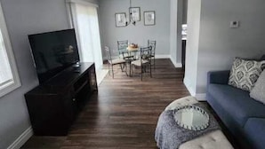Ein Wohnzimmer mit dunklem Holzboden, einem blauen Sofa, einem Fernseher und einem Essbereich mit Tisch und Stühlen.