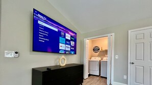 Habitación con televisor de pantalla plana montado en la pared mostrando la interfaz de un servicio de streaming, un aparador negro, una cesta de ropa y una puerta que da a otra habitación.
