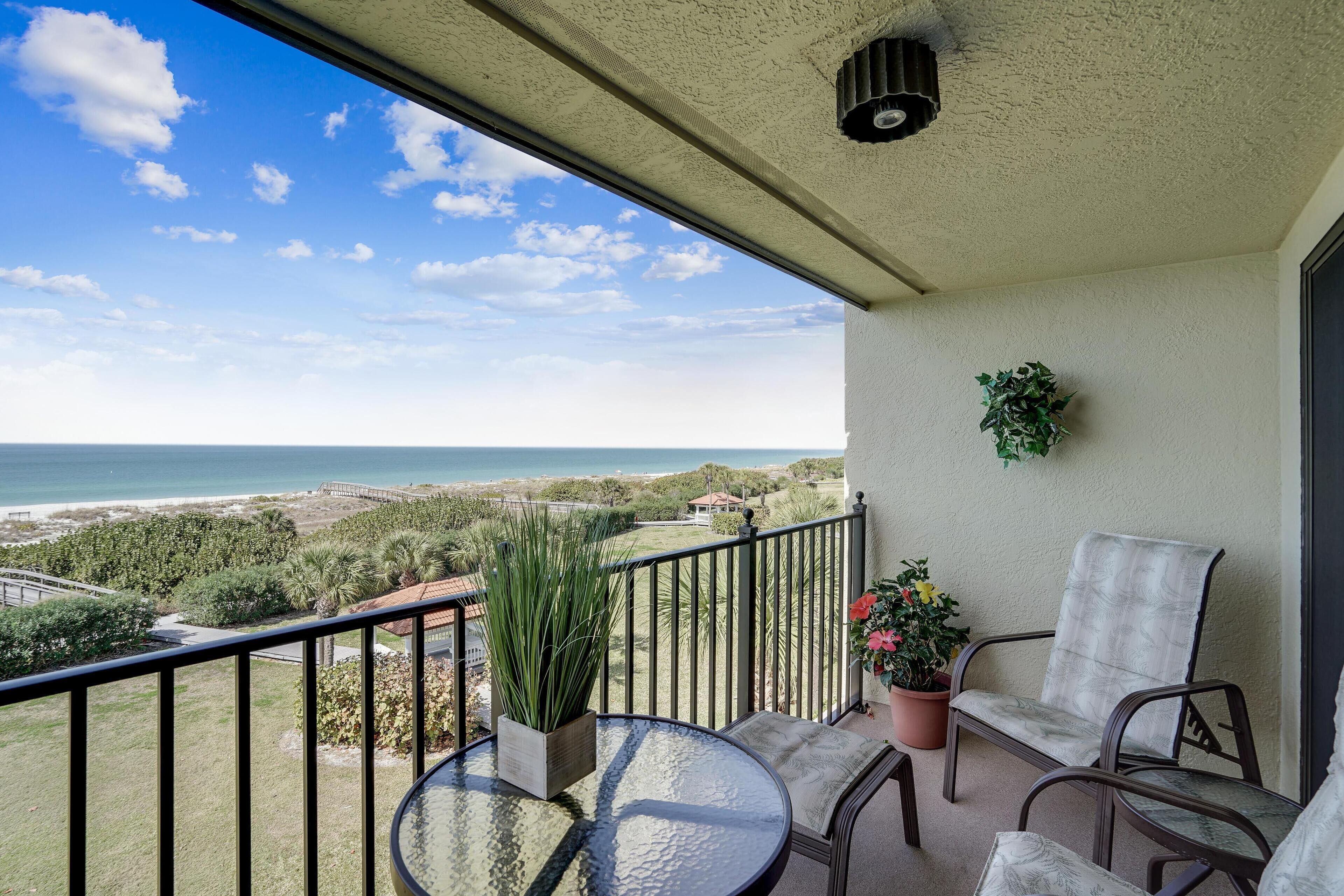 A balcony with a glass table, two chairs, a potted plant, and a view of the ocean and greenery.