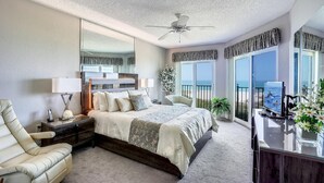 A bedroom with a large bed, a desk, a chair, a ceiling fan, and a view of the ocean. - Land's End 6-404 Beach Front (Treasure Island)