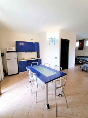 Een kleine keuken met blauwe kastjes, een witte koelkast en een eettafel met stoelen.