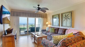 A living room with a sofa, a coffee table, a TV, and a balcony with a view of the ocean. - Land's End 7-304 Beach Front - Premier (Treasure Island)