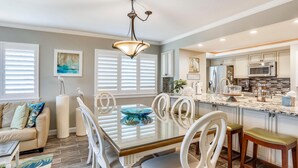 A modern kitchen with white cabinetry, a central island, and a dining area with a glass-top table and white chairs. - Land's End 7-306 Beach Front - Premier (Treasure Island)