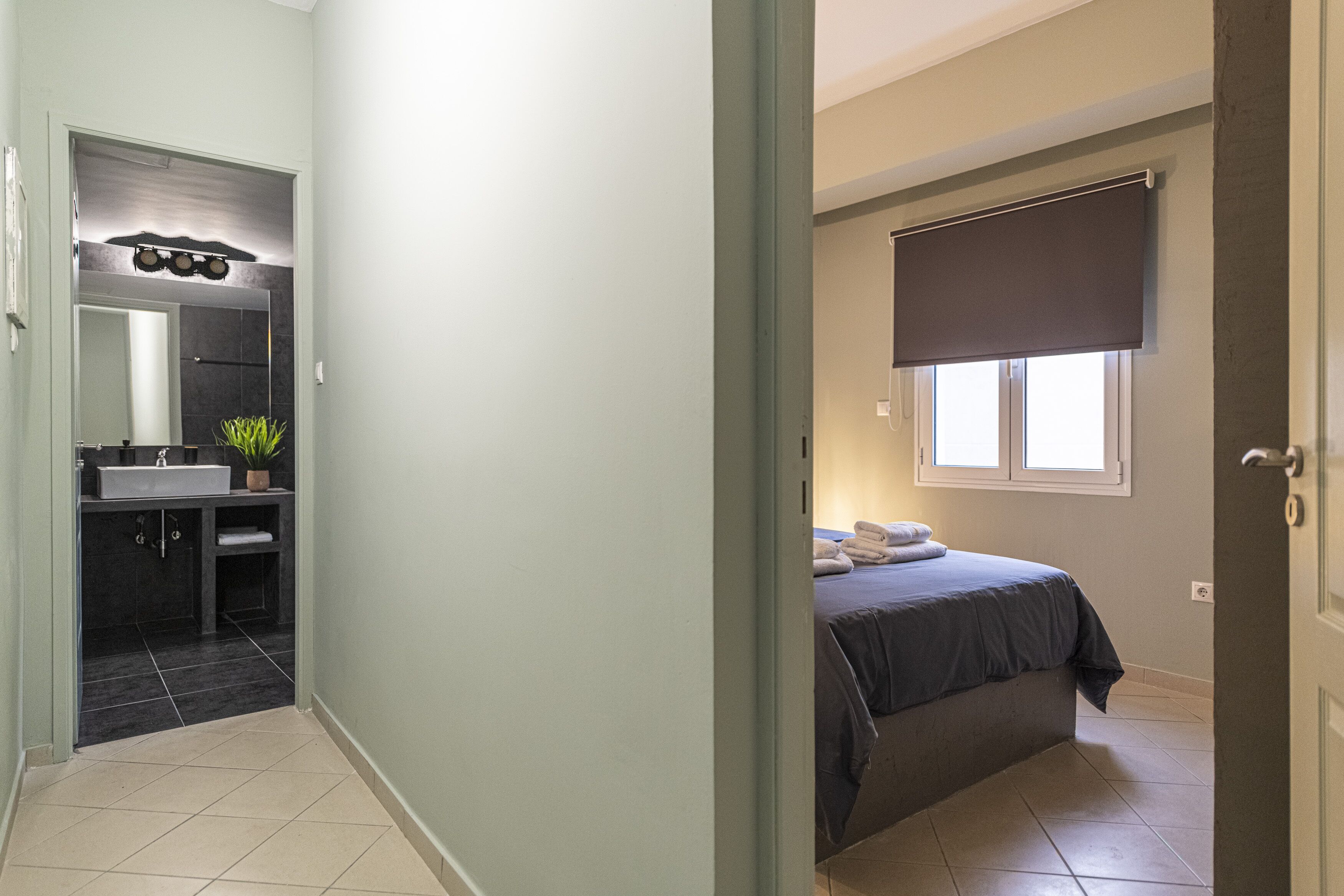 A bedroom with a bed, a window with a blind, and a bathroom with a sink and mirror.