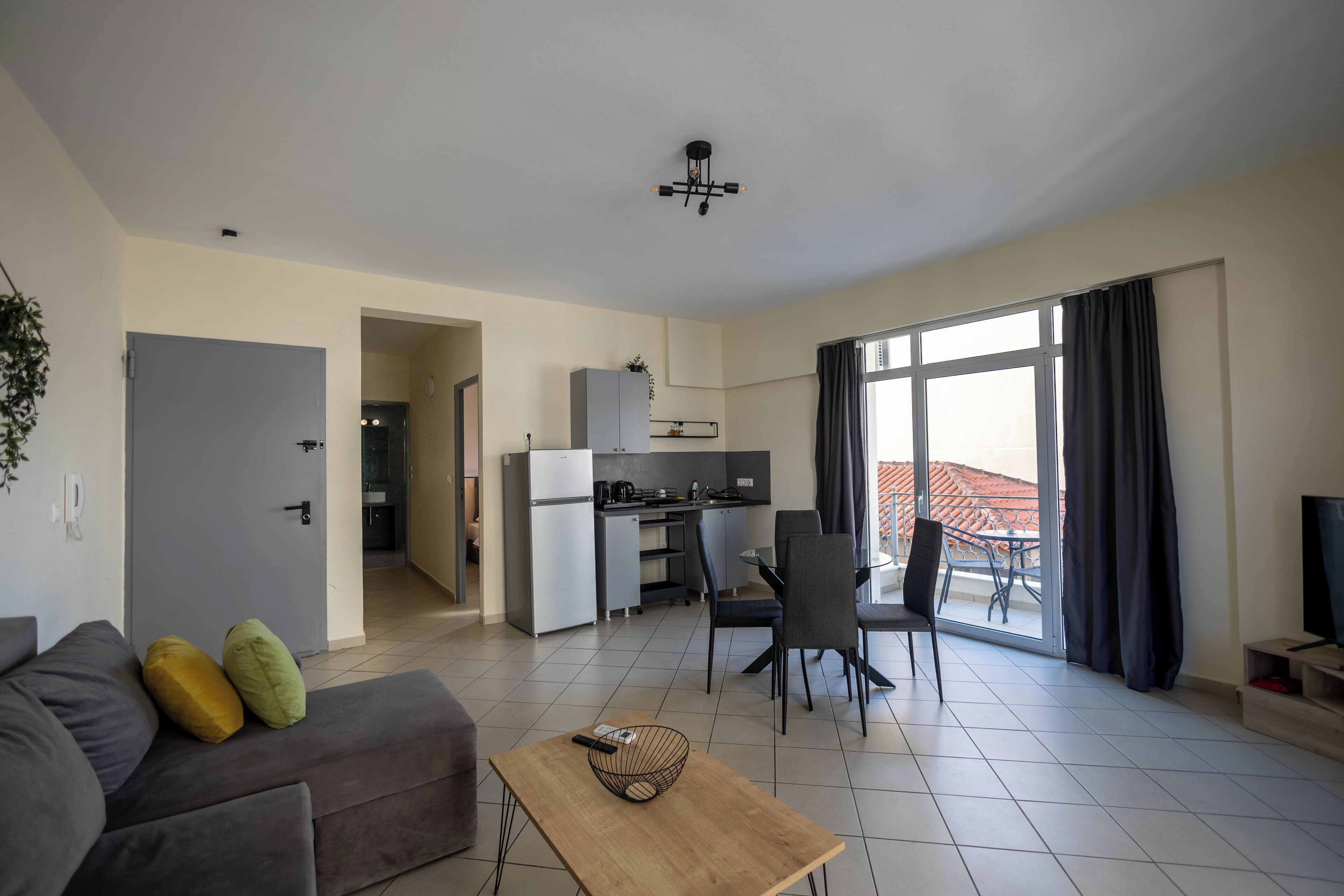 A modern living room with a sofa, a coffee table, and a dining area with chairs. There is a kitchen in the background, and a balcony with a view.