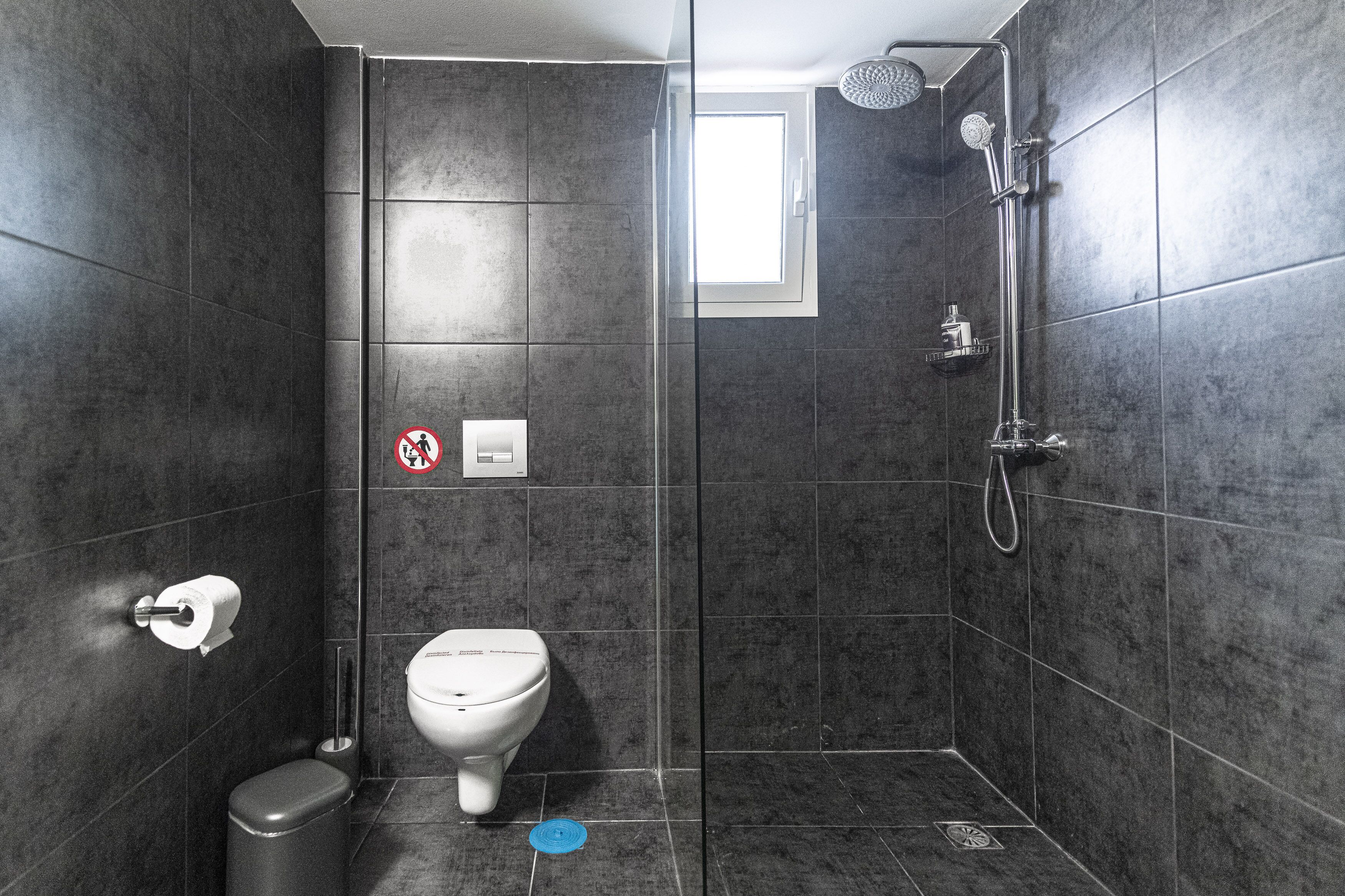 Egy modern fürőszoba sötét színű csempékkel, WC-vel, zuhanyzóval és egy „belépés tilos” táblával.