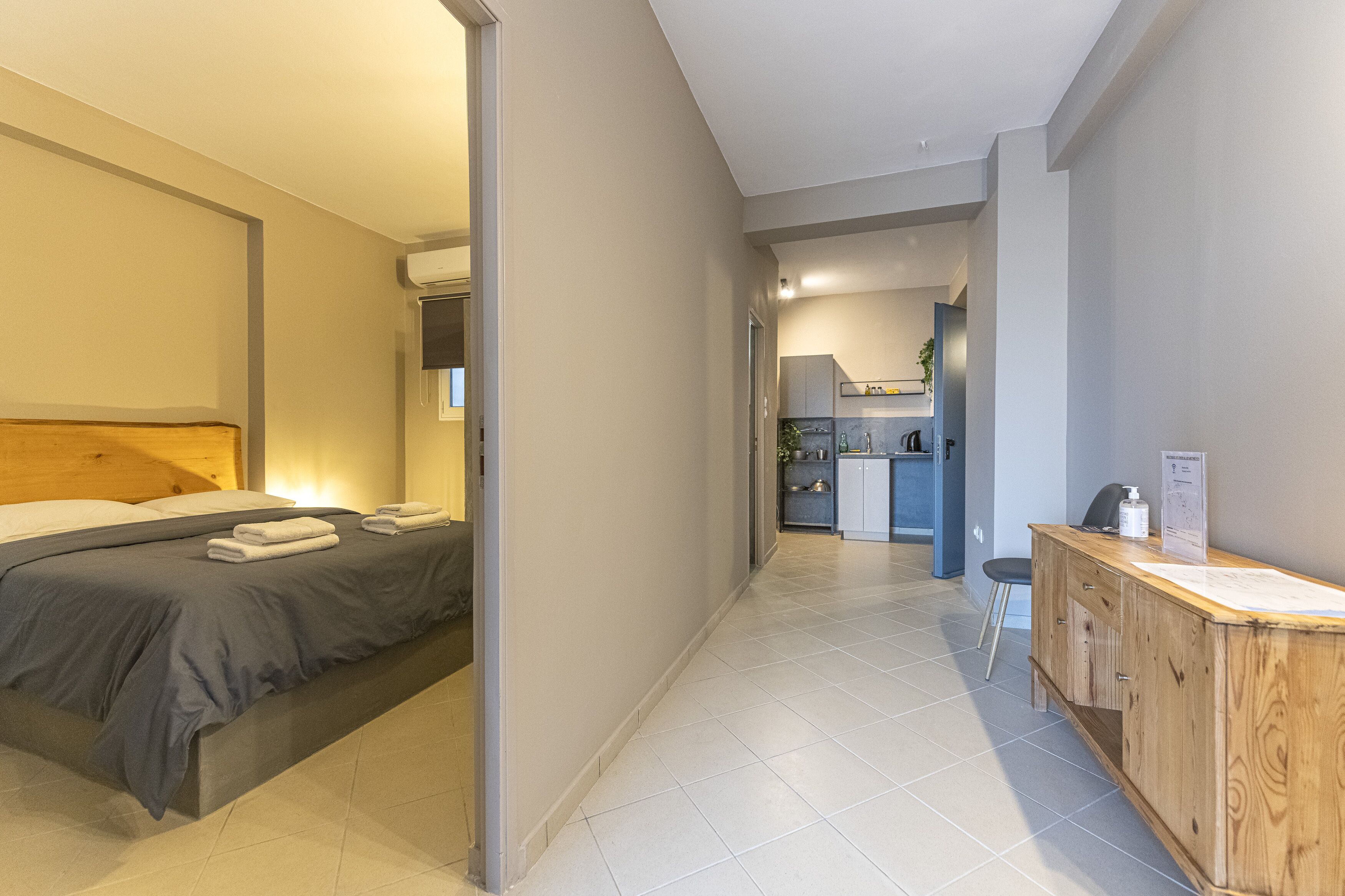 A modern hotel room with a neatly made bed, a wooden desk with a chair, and a kitchenette in the background.
