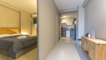 A modern hotel room with a neatly made bed, a wooden desk with a chair, and a kitchenette in the background.