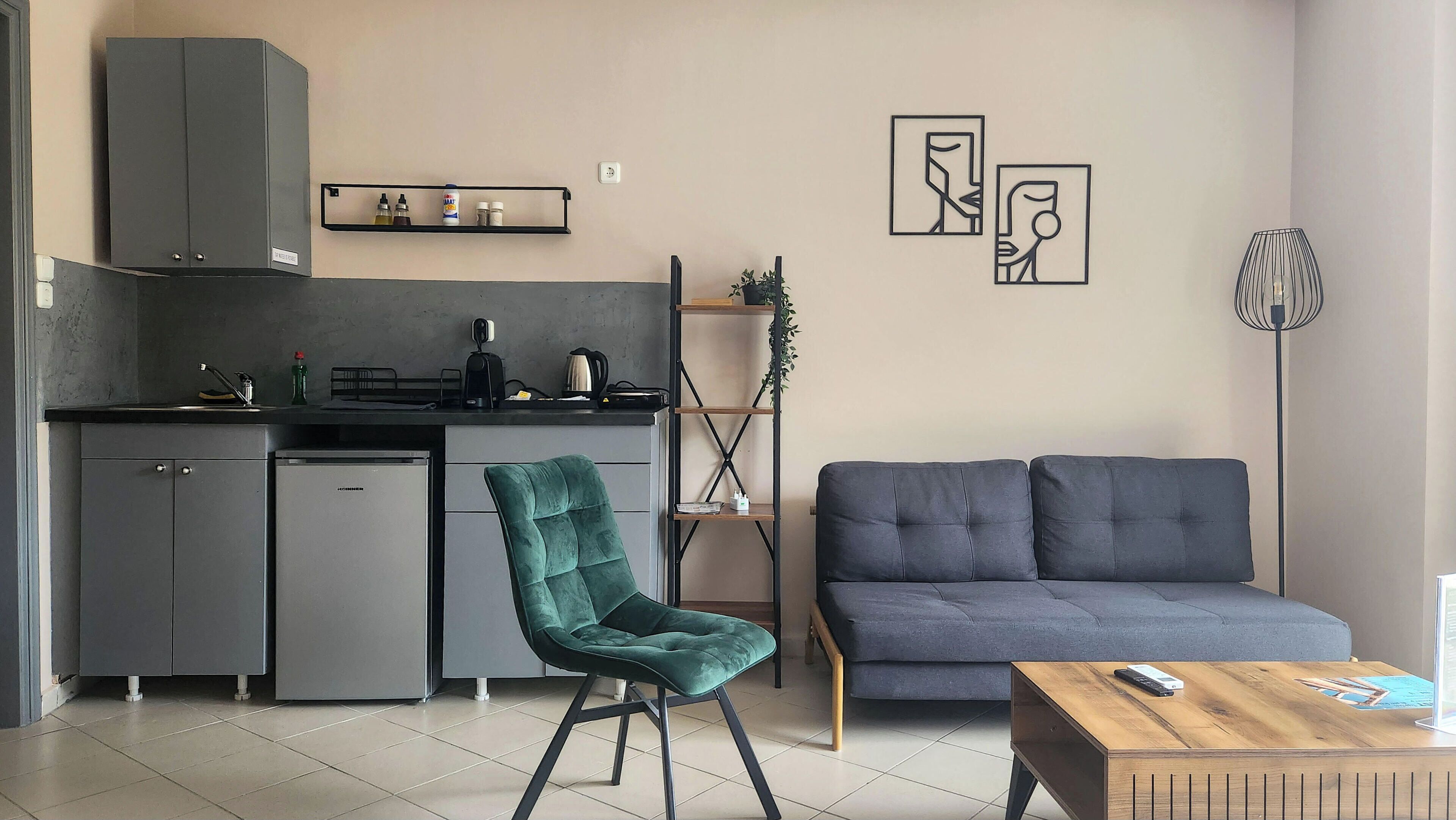 A modern kitchen with a built-in refrigerator, a coffee machine, and a sink. There is a teal chair and a gray sofa in the living area, with a wooden coffee table in front.