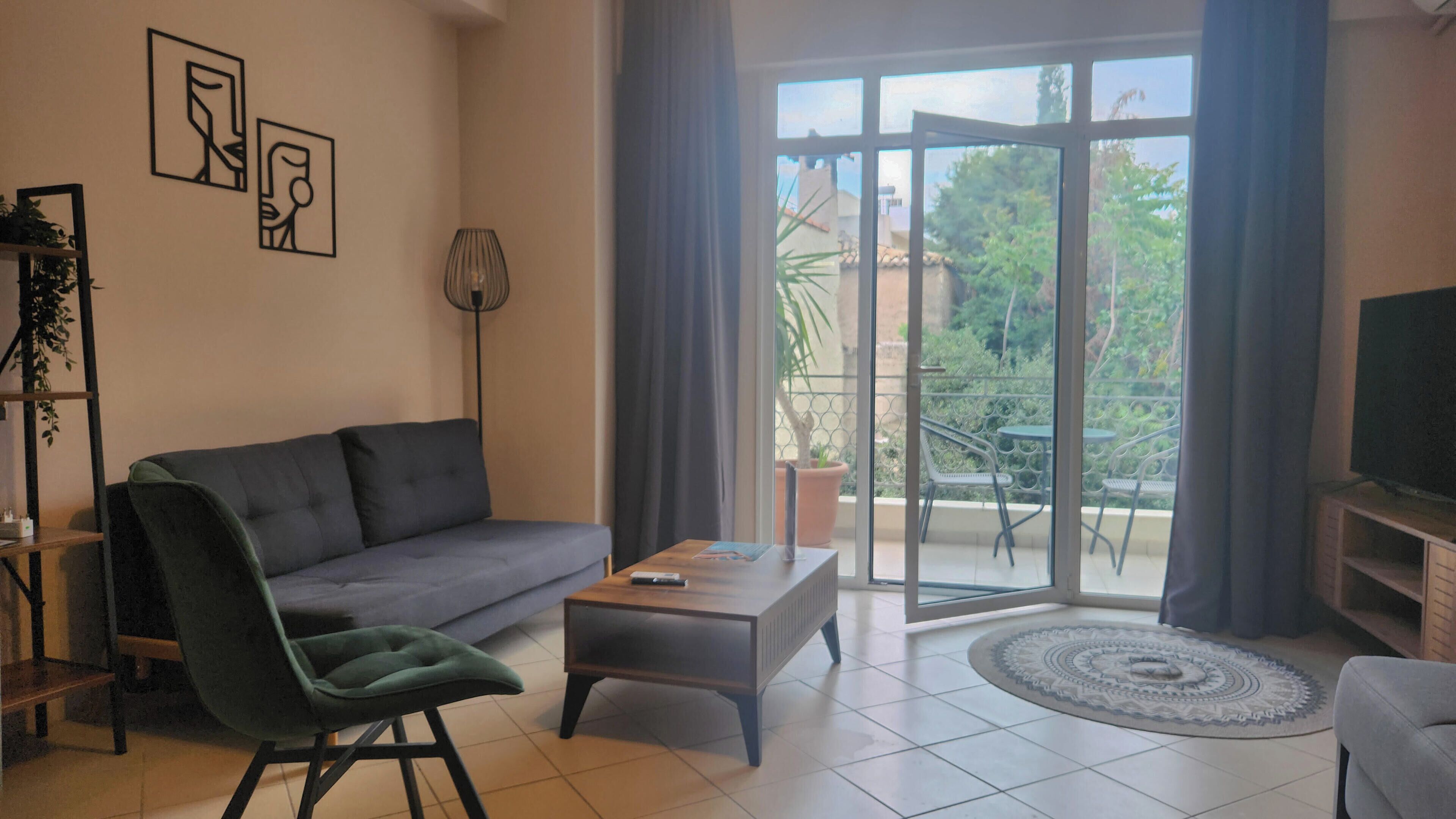 Un salon avec un canapé gris, un fauteuil vert, une table basse et un téléviseur posé sur un meuble. Il y a un balcon avec vue sur la verdure.