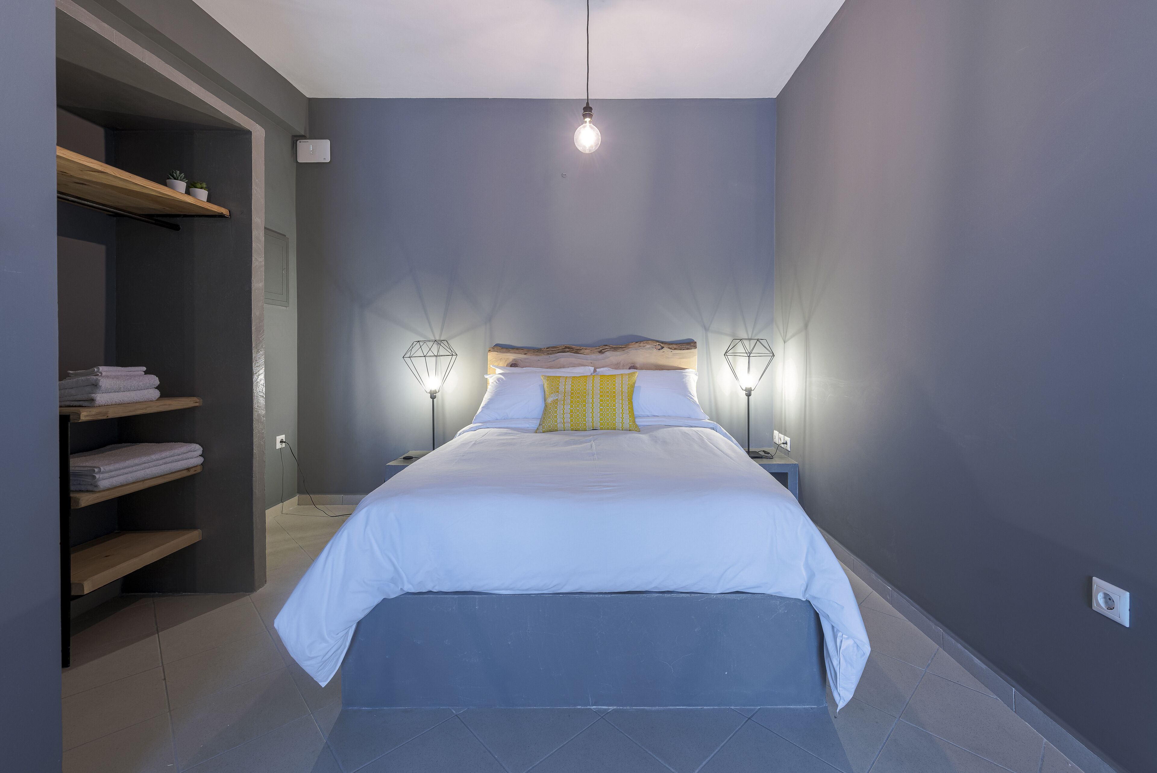 A bedroom with a bed, bedside lamps, a shelf with towels, and a hanging light fixture.