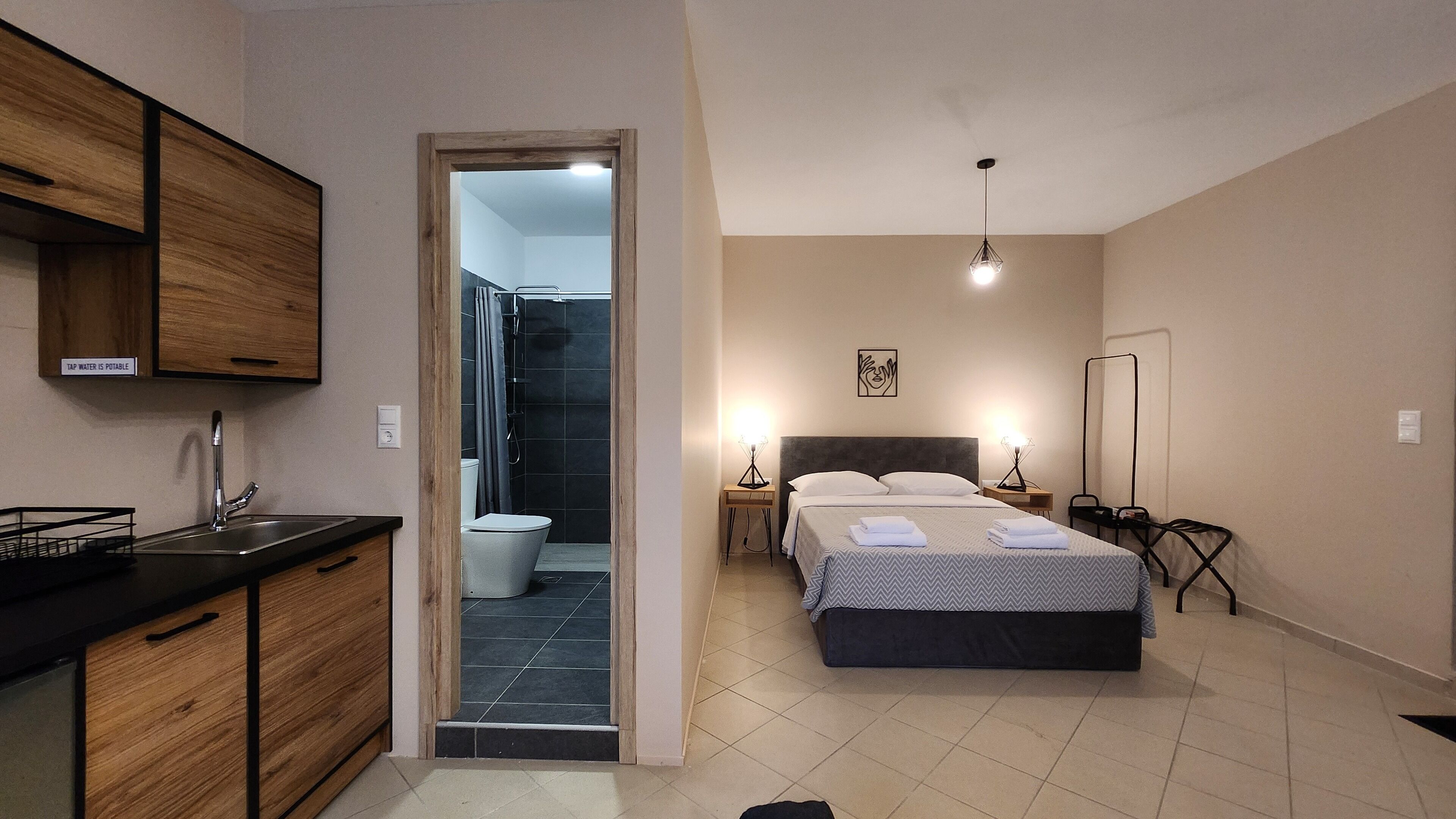 A compact hotel room with a kitchenette, a shower, a bed with a headboard, bedside lamps, and a framed picture on the wall.