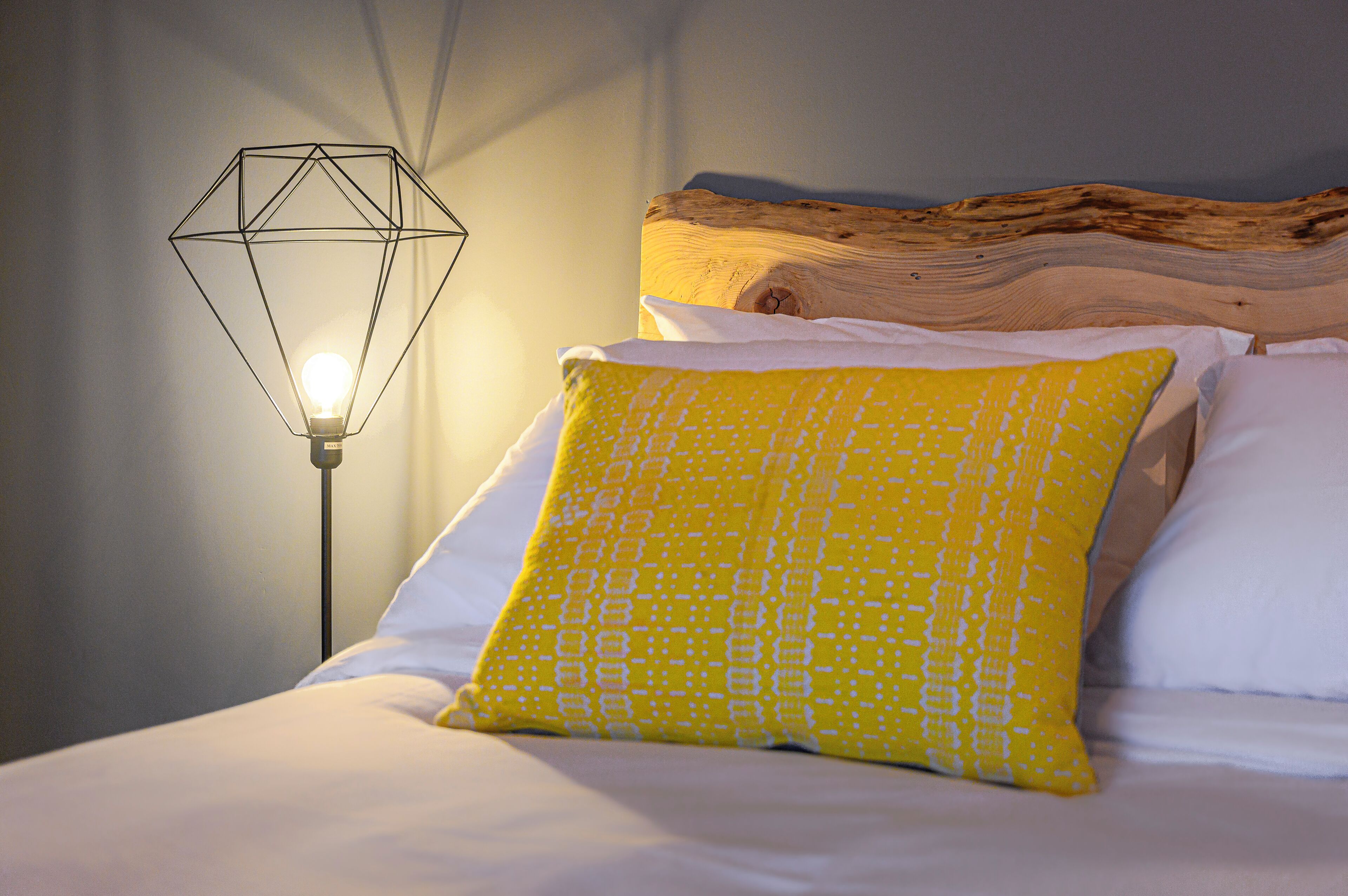 A bed with white and yellow pillows, a wooden headboard, and a geometric light fixture.