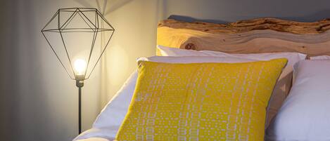 Una cama con almohadas blancas y amarillas, un cabecero de madera y una lámpara con forma geométrica.