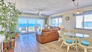 A spacious living area with a brown leather sofa, a glass-top dining table surrounded by white wicker chairs with blue cushions, and a bamboo plant in a woven pot. - Land's End 7-406 Beach Front - Premier (Treasure Island)