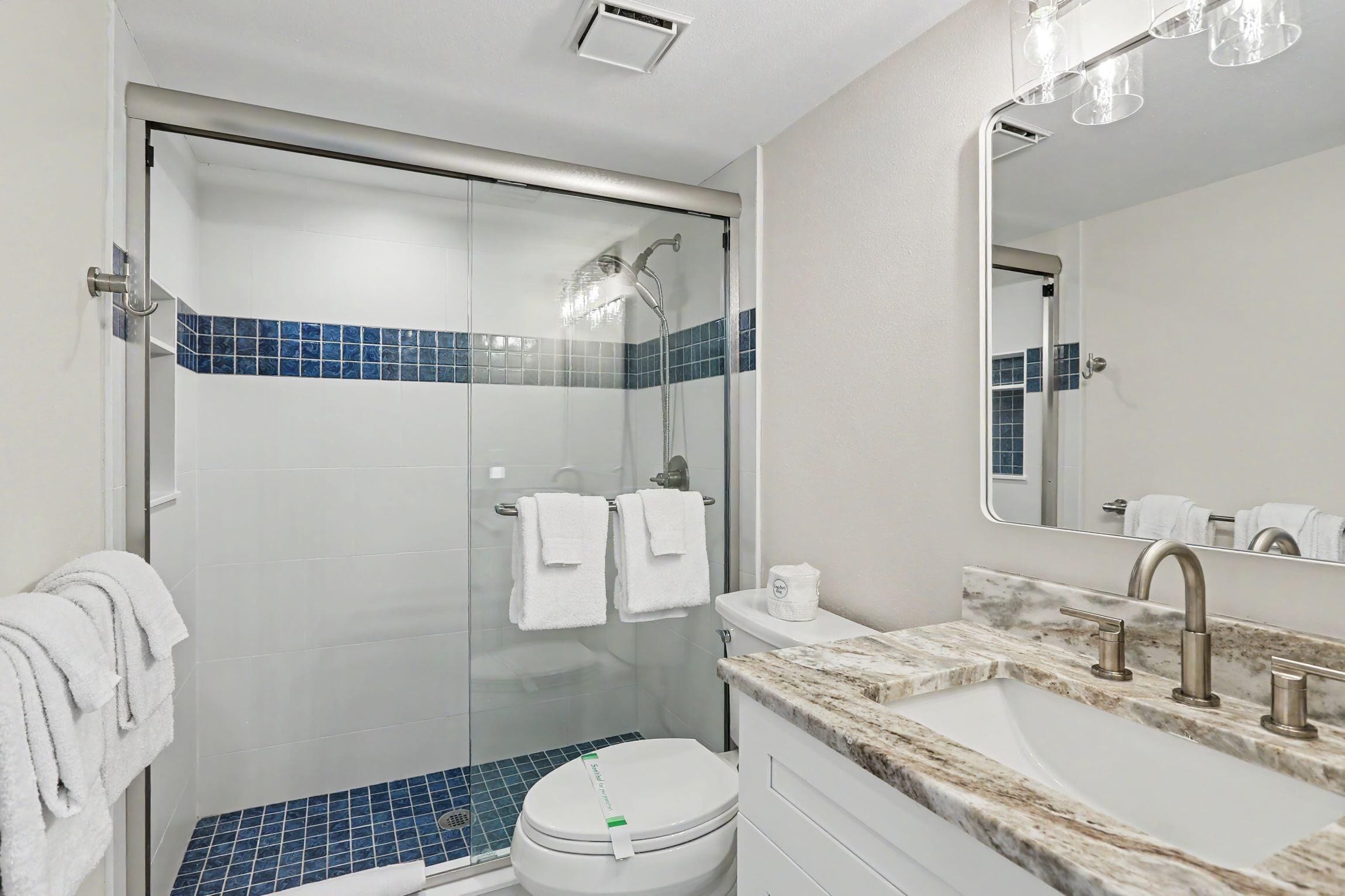 A bathroom with a glass-enclosed shower, a white toilet, a sink with a marble countertop, and a mirror above the sink.