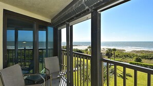 A balcony with a glass door leading to a patio area, a table with chairs, and a view of the beach. - Land's End 8-403 Beach Front - Premier (Treasure Island)