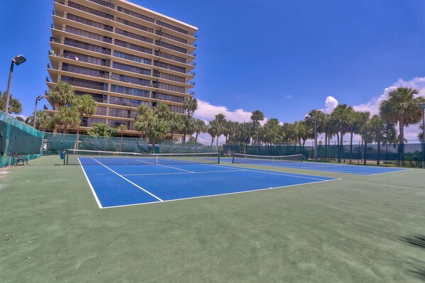 Cancha de tenis al aire libre con red, rodeada de palmeras y un edificio de varios pisos al fondo.