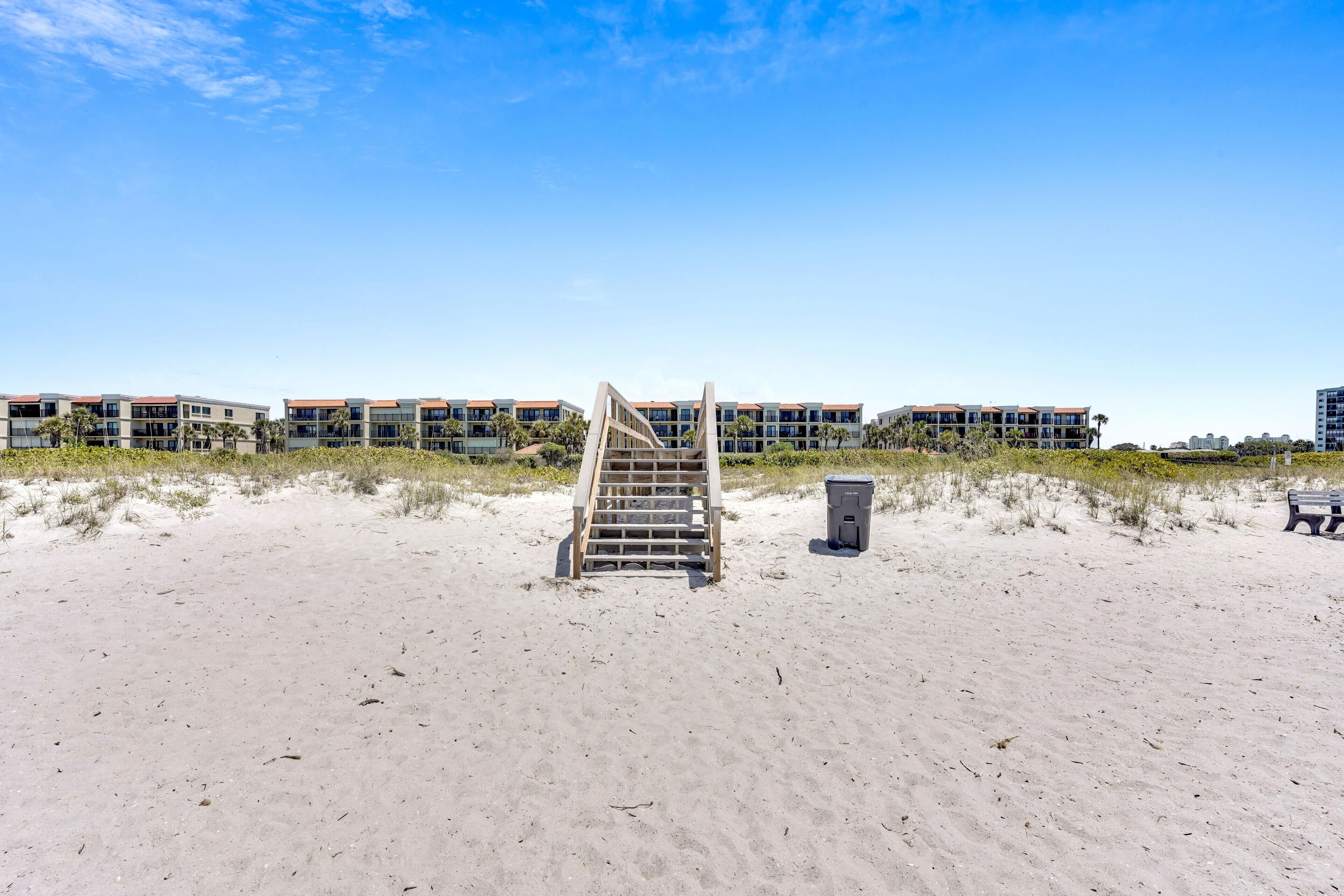 A beach with a wooden staircase leading to a building with multiple balconies, surrounded by sand and sparse vegetation.