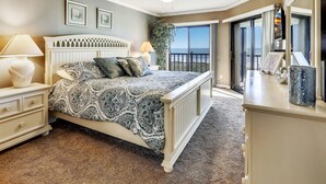 Une chambre avec un grand lit, une table de chevet, une commode et un ventilateur de plafond. Une grande fenêtre offre une vue sur l’océan.