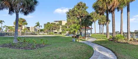 A paved pathway curving through a well-maintained grassy area with palm trees and a building in the background.