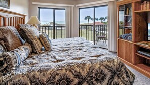 Een slaapkamer met een groot bed, een nachtkastje, een lamp, een boekenkast en een balkon met uitzicht op het strand en palmbomen.