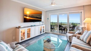 A living room with a large TV, a glass coffee table, striped sofas, a cabinet with glass doors, and a balcony with outdoor seating. - Land's End 9-304 Beach Front - Premier (Treasure Island)