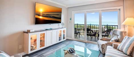 A living room with a large TV, a glass coffee table, striped sofas, a cabinet with glass doors, and a balcony with outdoor seating.
