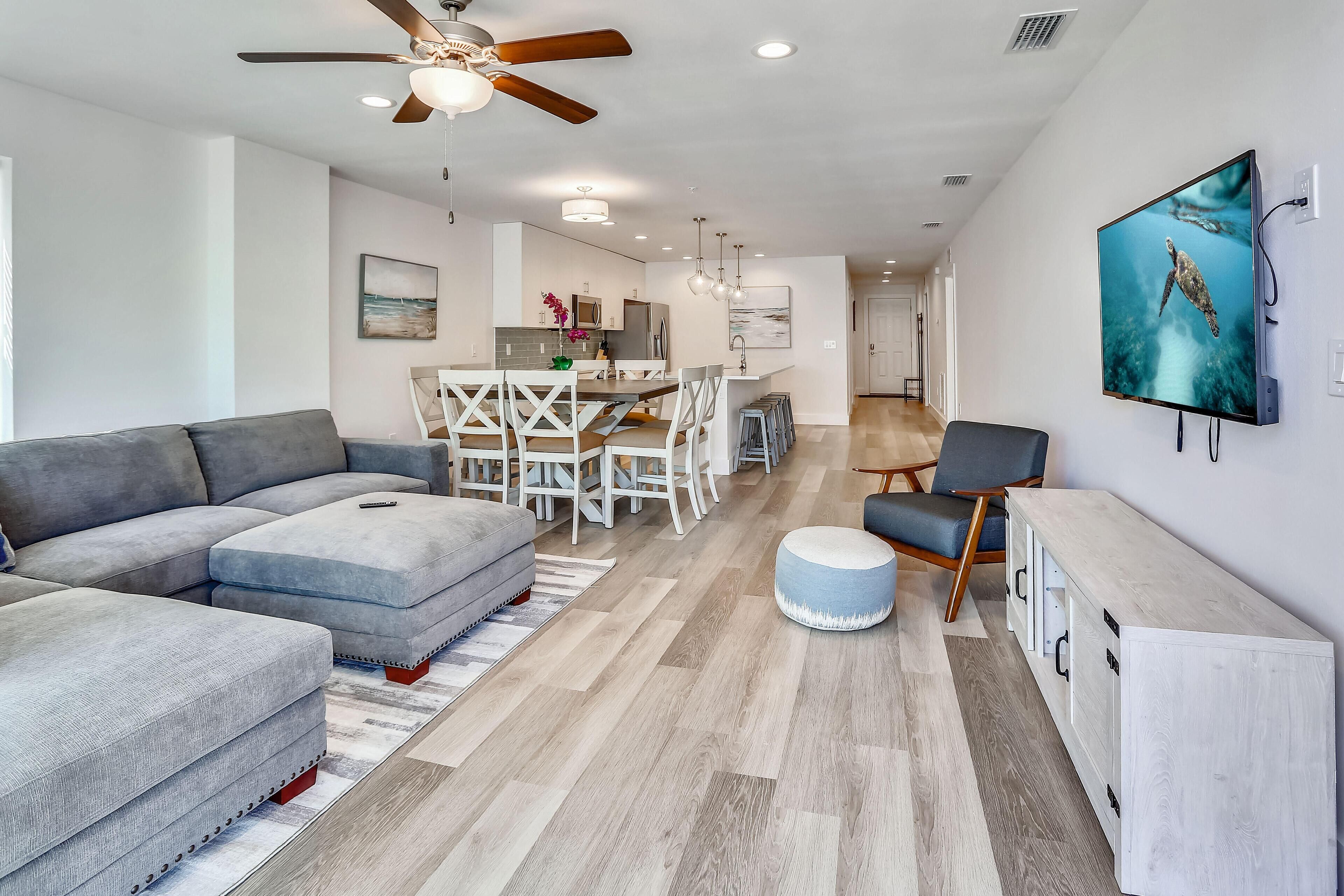 A modern living room with a gray sectional sofa, a dining area with a white table and chairs, and a kitchen in the background.