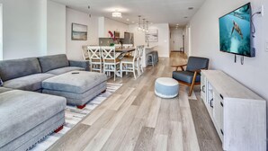 Un salon moderne avec un canapé d’angle gris, un coin repas avec une table et des chaises blanches, et une cuisine en arrière-plan.