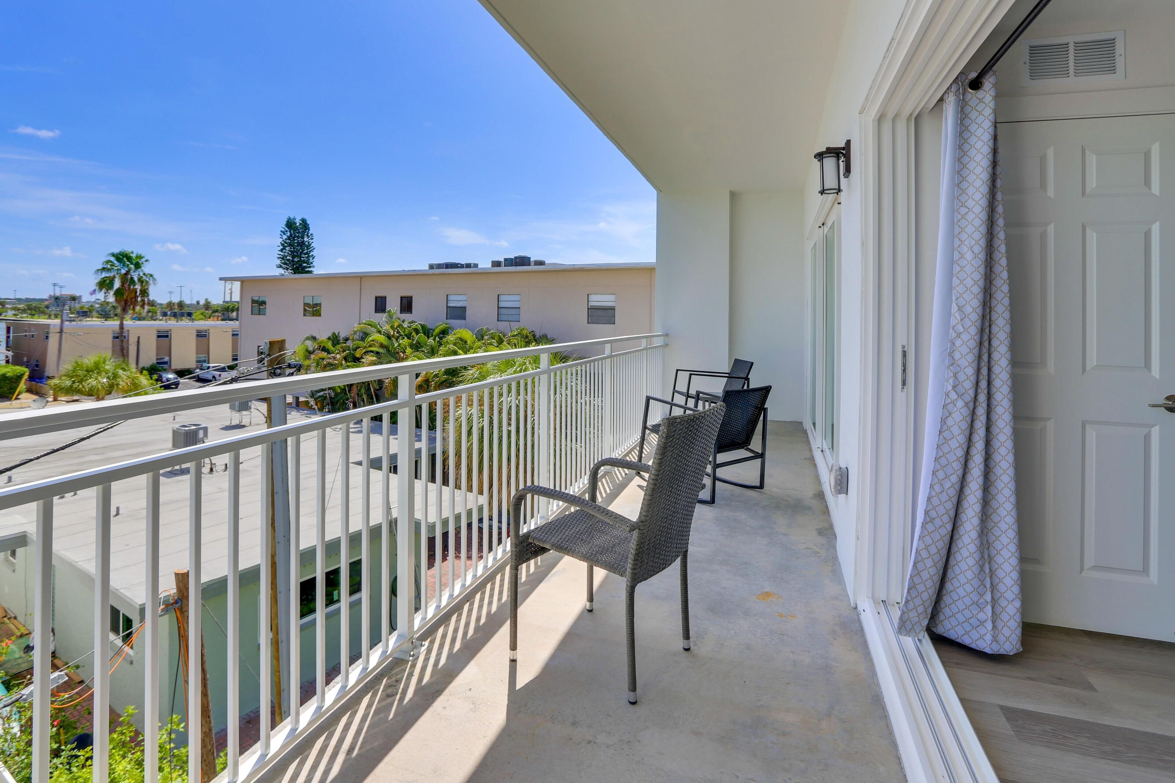 Ein Balkon mit weißer Brüstung, einem Stuhl und einem Tisch. Eine Tür mit gemustertem Vorhang und ein Gebäude im Hintergrund.