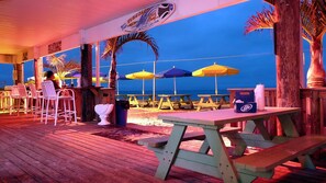 A beachside restaurant with outdoor seating, palm trees, and a view of the ocean.