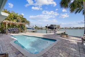 A swimming pool with a pergola and outdoor seating area, surrounded by palm trees and a view of a marina.