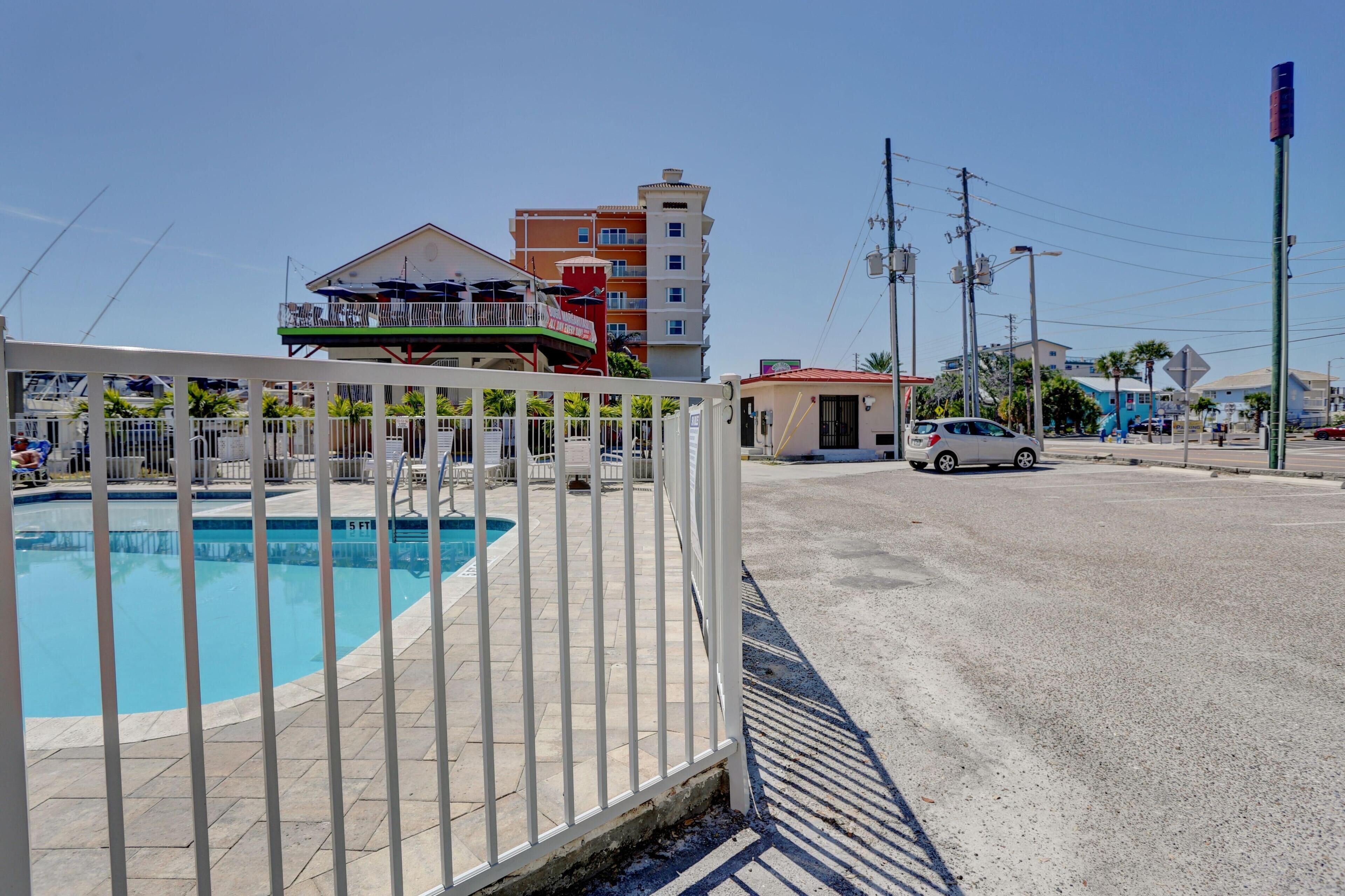 A swimming pool with a white fence, a white car parked on the street, and buildings in the background.