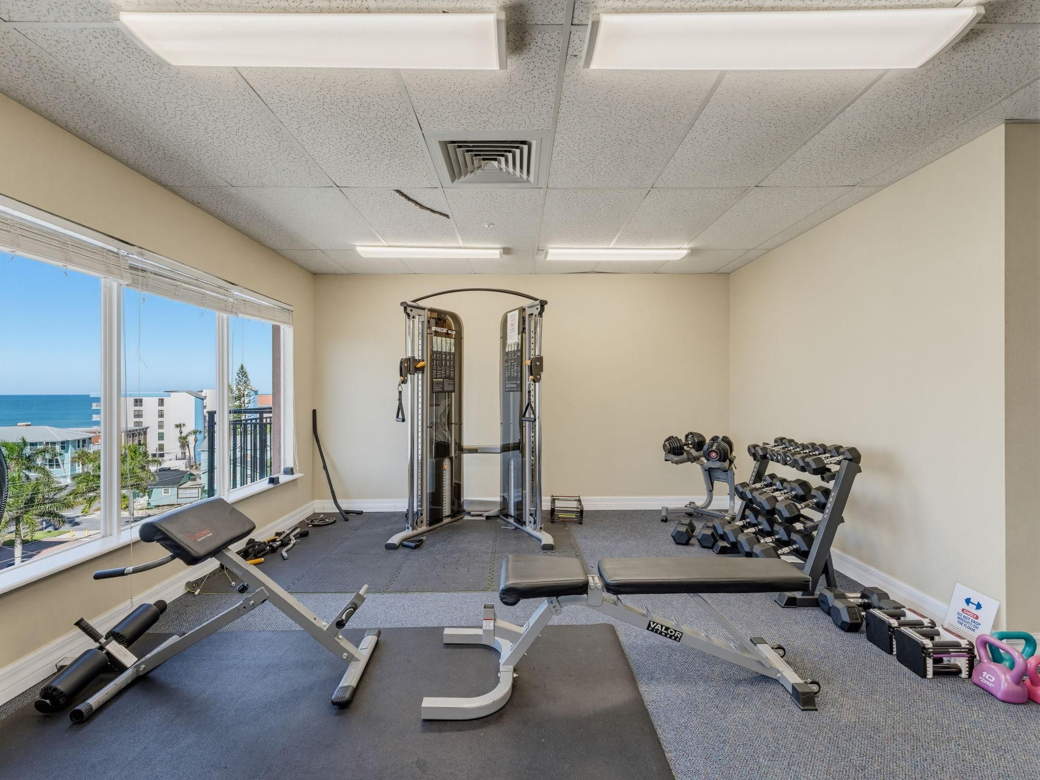Une salle de sport équipée de matériel d’exercice, une fenêtre donnant sur des immeubles et un plafond avec des luminaires.