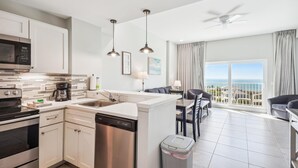 A modern kitchen with white cabinets, a dishwasher, and a coffee maker. There is a dining area with a table and chairs, and a view of the ocean through large windows. - Madeira Bay Resort I 1504 (Madeira Beach)