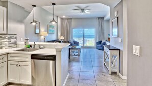 A modern kitchen with a dishwasher, a dining area with a table and chairs, and a view of the ocean through large windows. - Madeira Bay Resort I 1504 (Madeira Beach)