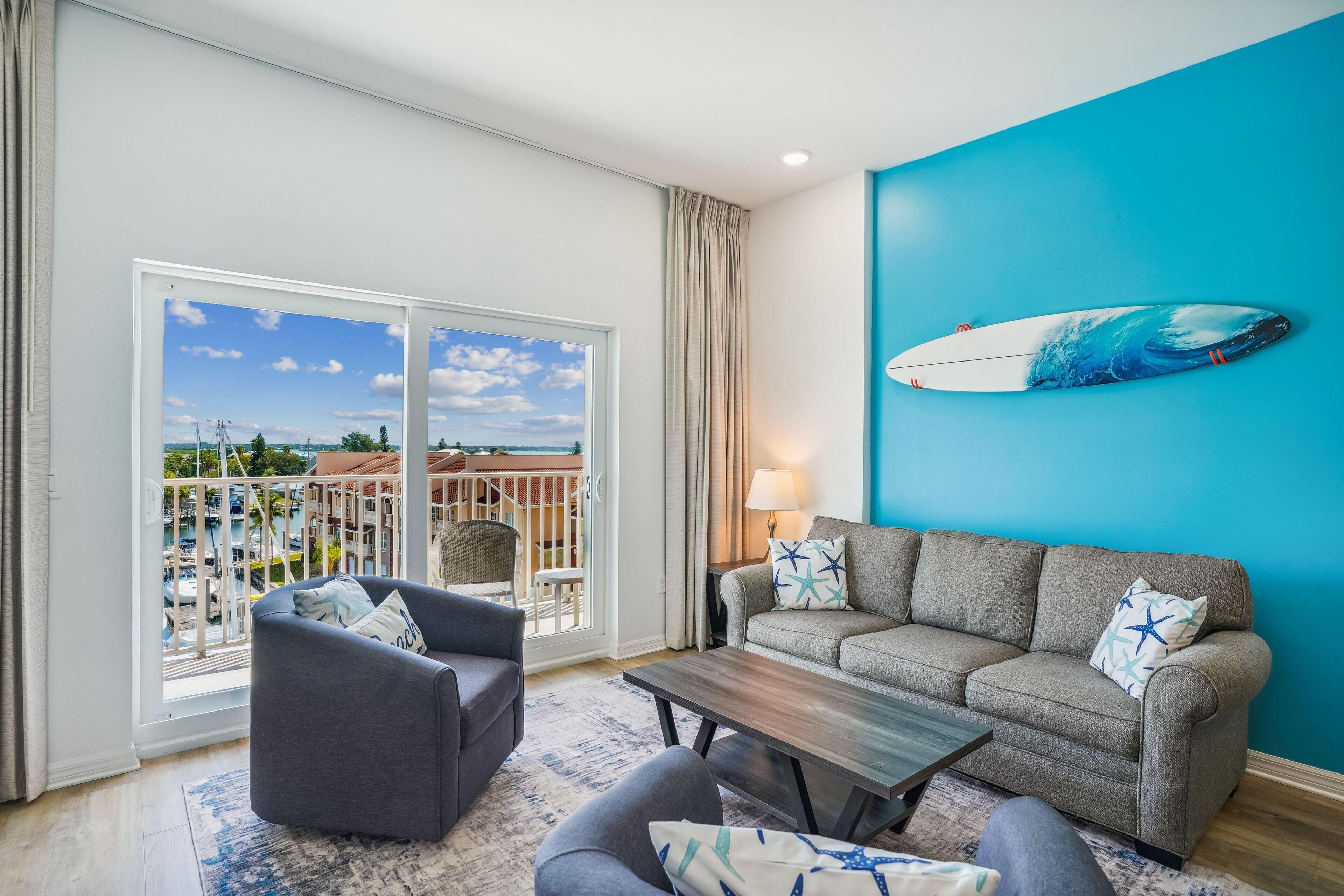 A living room with a grey sofa, a smaller grey armchair, a wooden coffee table, and a blue wall with a surfboard decoration.