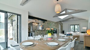 Un comedor con una mesa de madera dispuesta para seis personas, rodeada de sillas blancas. Se aprecia una pared de piedra y un ventanal grande al fondo.