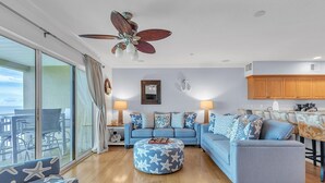 Un salon avec un canapé bleu, un pouf rond orné de motifs en étoile, un ventilateur de plafond et une vue sur l’extérieur, accessible par une porte-fenêtre coulissante.