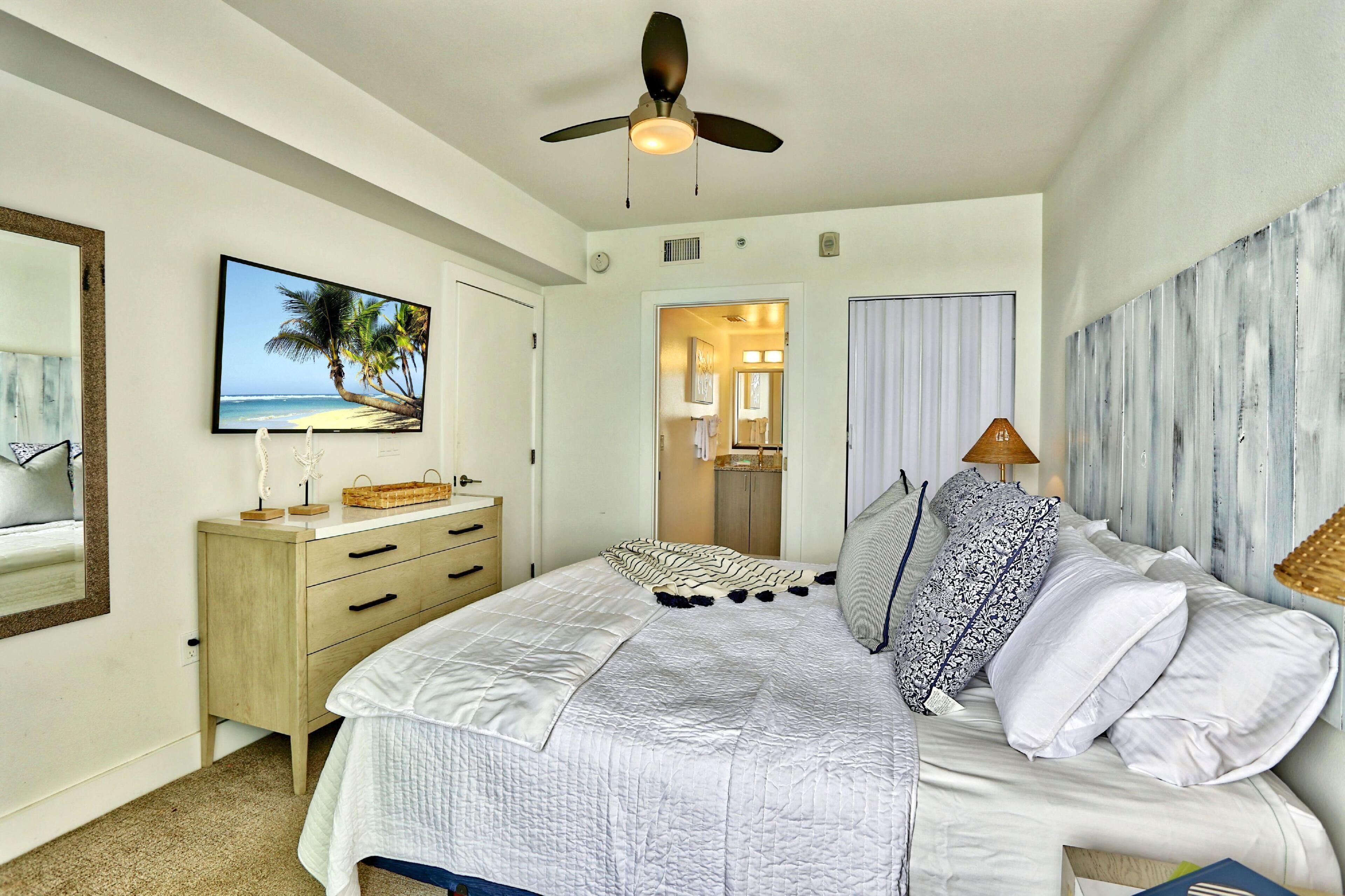 A bedroom with a bed, a ceiling fan, a painting of a beach, a dresser, and a door leading to a bathroom.