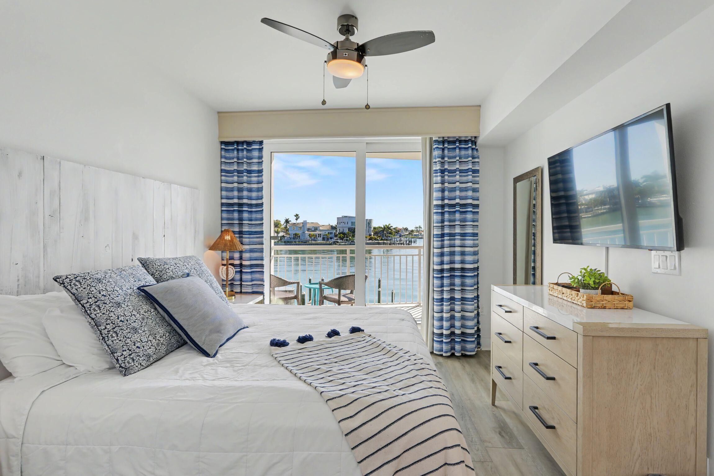 Un dormitorio con cama, ventilador de techo, televisor, balcón con vistas y cómoda.