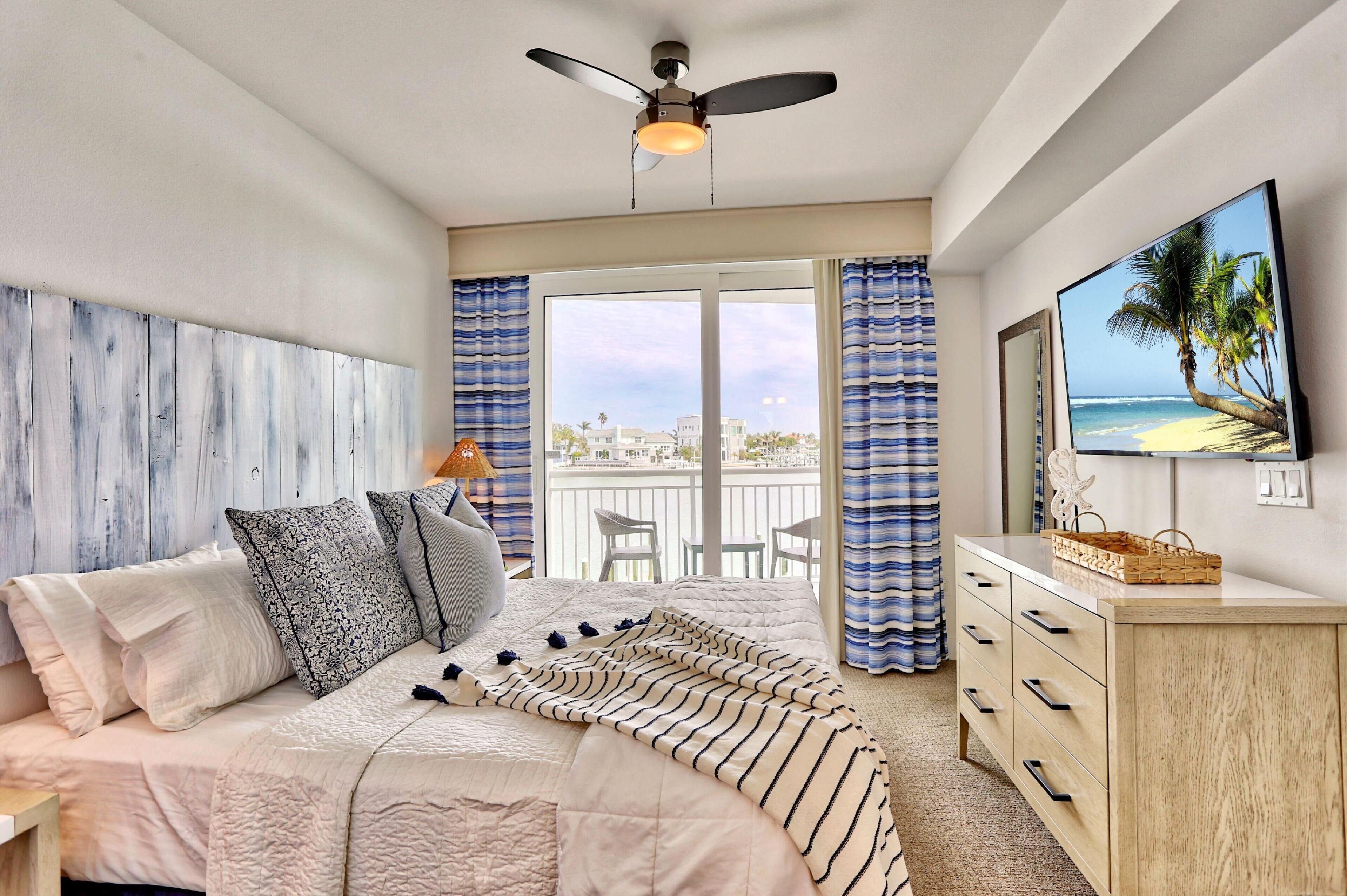A bedroom with a bed, a ceiling fan, a balcony with a view, and a TV on the wall.