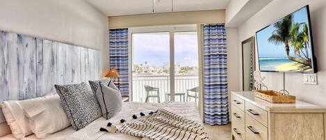 A bedroom with a bed, a ceiling fan, a balcony with a view, and a TV on the wall.