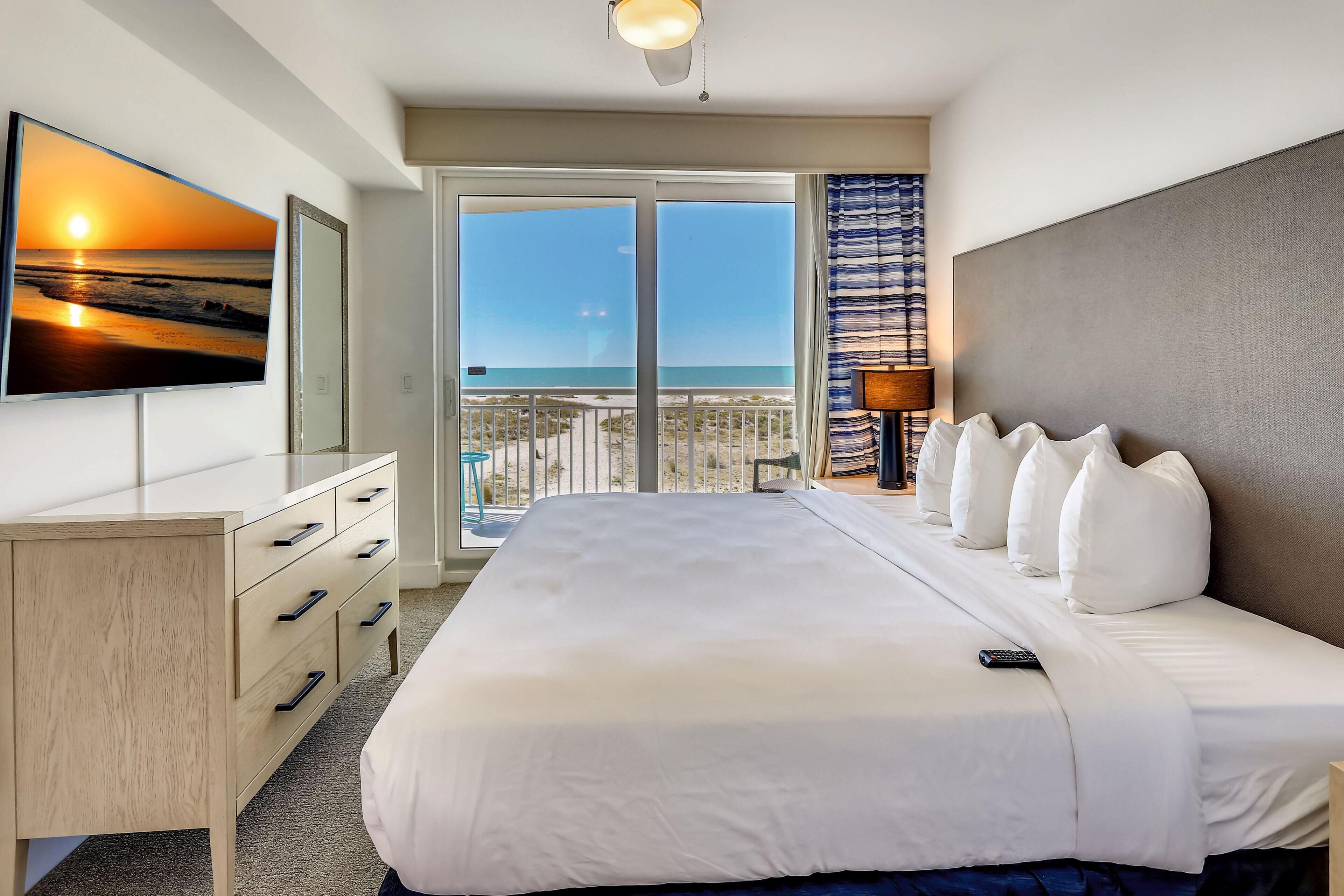 A hotel room with a bed, a desk, a TV, and a balcony with a view of the sea.